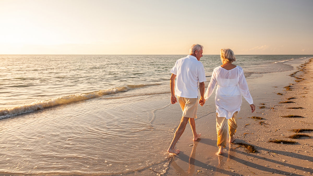 Happy man and senior woman walking and holding hands on the beach at sunset